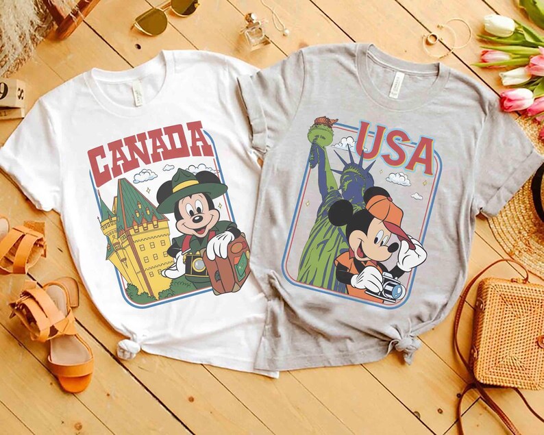 Pode incluir: Duas camisetas com gr&aacute;ficos tem&aacute;ticos de viagem. Uma &eacute; branca com "CANADA" e um rato de desenho animado com um chap&eacute;u verde. A outra &eacute; cinza com "USA" e um rato de desenho animado com um bon&eacute; laranja, com a Est&aacute;tua da Liberdade.