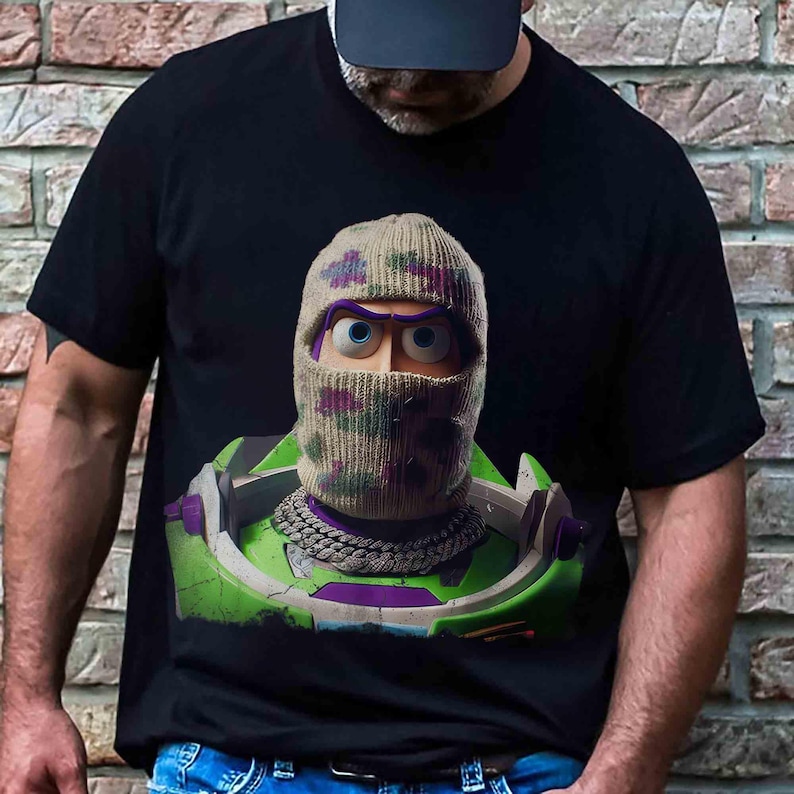 May include: A black t-shirt featuring a graphic print of a character wearing a camouflage-patterned knit balaclava and a thick silver chain necklace. The character is depicted in a green and purple space-themed suit with a high collar design.