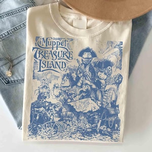 Może przedstawiać: Kremowy t-shirt z niebieskim nadrukiem graficznym Muppet Treasure Island. Wzór przedstawia postacie w strojach piratów, statek, mapę skarbów oraz skrzynię wypełnioną złotymi monetami. Napis Muppet Treasure Island jest wydrukowany stylizowaną czcionką po lewej stronie grafiki.