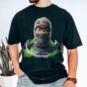 May include: A black t-shirt featuring a graphic print of a character wearing a camouflage-patterned knit balaclava and a silver chain necklace. The character is encased in a green, purple, and white space-themed suit with a high collar.