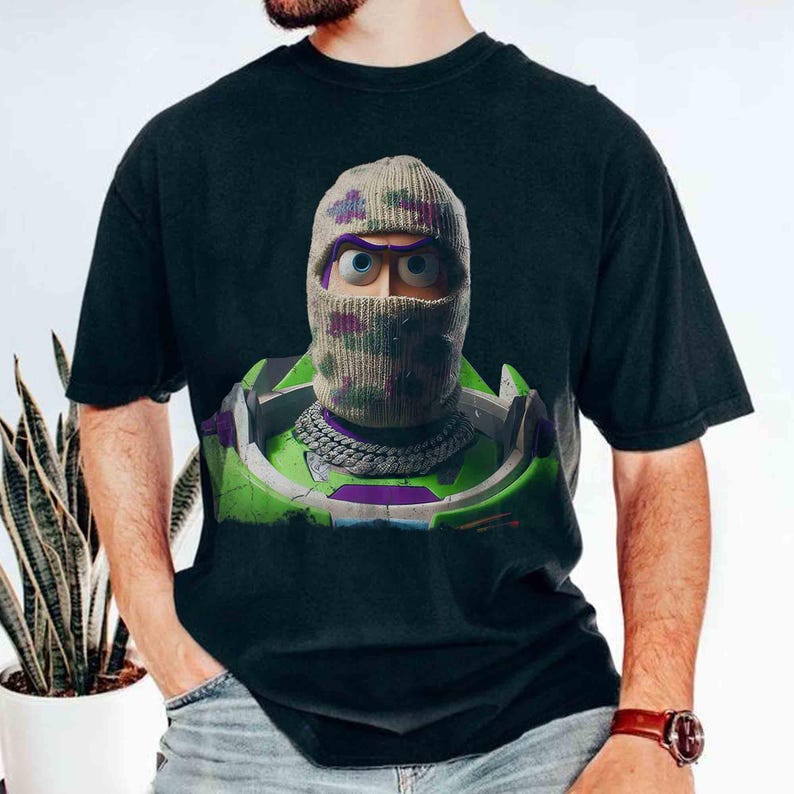 May include: A black t-shirt featuring a graphic print of a character wearing a camouflage-patterned knit balaclava and a silver chain necklace. The character is encased in a green, purple, and white space-themed suit with a high collar.