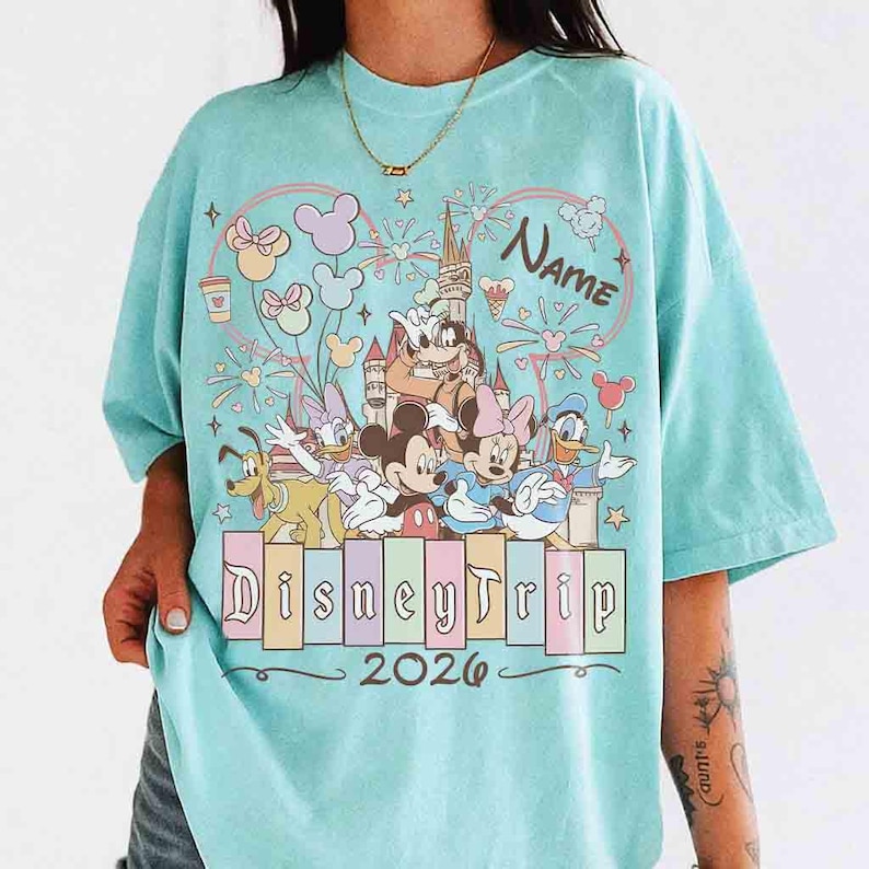 K&ouml;nnte beinhalten: Hellblaues T-Shirt mit einem farbenfrohen Disney-Motiv. Das Design zeigt Micky und Minnie Maus, Donald Duck und andere Charaktere, mit dem Text "DisneyTrip 2026" und Platz f&uuml;r einen Namen. Das Shirt hat eine lockere Passform.