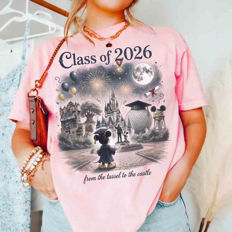 May include: Pink t-shirt with a graphic featuring Mickey Mouse in a graduation gown, standing in front of a castle and other landmarks. The text "Class of 2026" and "from the tassel to the castle" are printed on the shirt.