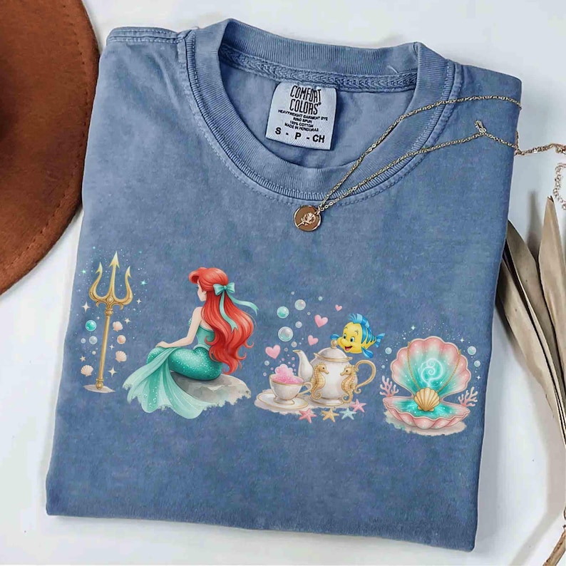 May include: Blue Comfort Colors t-shirt featuring a colorful graphic of Ariel from The Little Mermaid. The design includes Ariel, Flounder, a seashell, and a teapot. A gold necklace with a pendant is draped on the shirt.