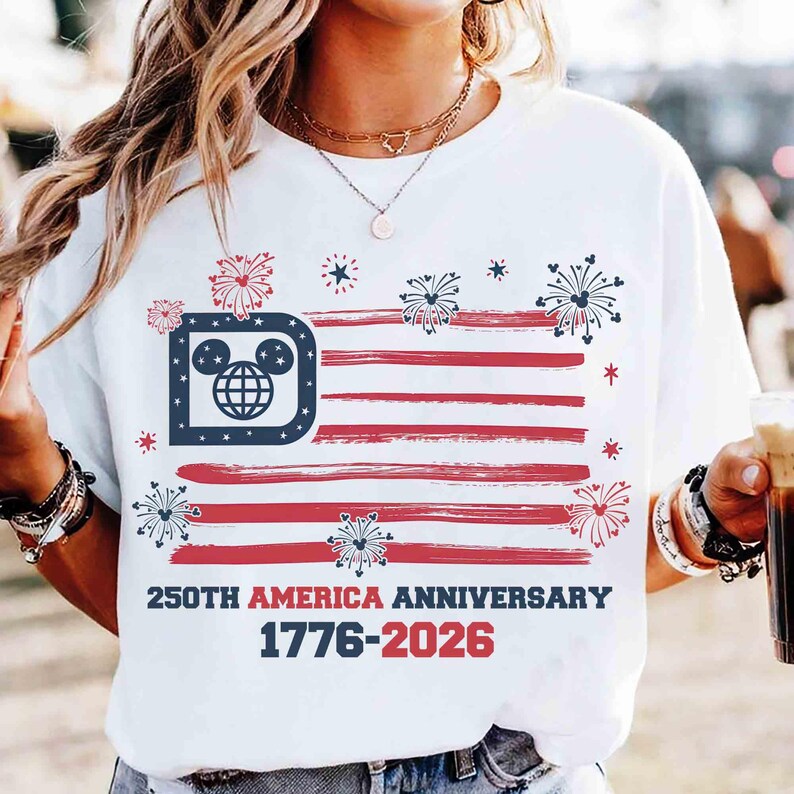 May include: White t-shirt featuring a stylized American flag design with red stripes and a blue corner with a Mickey Mouse head logo. The text "250th AMERICA ANNIVERSARY 1776-2026" is printed below the flag design.