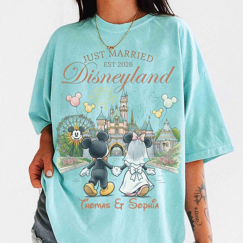 Może przedstawiać: Jasnoniebieska koszulka z motywem ślubu w Disneylandzie. Grafika przedstawia Myszki Miki i Minnie w strojach ślubnych, trzymające się za ręce przed zamkiem. Tekst zawiera "Just Married Disneyland Est 2026" i imiona.