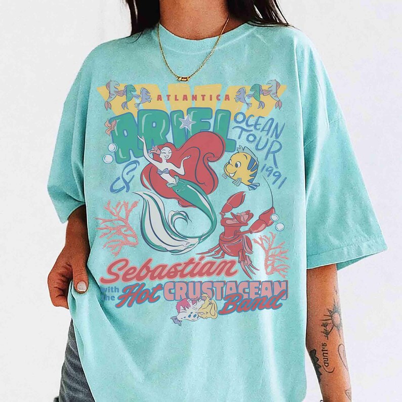 May include: A light blue t-shirt featuring a colorful graphic of Ariel from The Little Mermaid. The design includes Ariel, Flounder, Sebastian, and the text "Ocean Tour 1991" and "Sebastian with the Hot Crustacean Band."