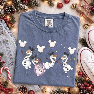May include: A blue crew neck t-shirt featuring a graphic of four Olaf snowmen characters, each wearing black mouse ears and holding a white mouse-shaped balloon. The shirt has a Comfort Colors brand label on the inside collar and small yellow stars.