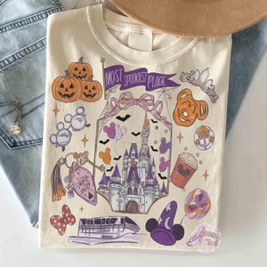 Pode incluir: Uma t-shirt cor de creme com uma impressão gráfica de um castelo, abóboras, um monotrilho e vários ícones. O texto "MOST SPOOKIEST PLACE" está numa faixa roxa. O design inclui morcegos, uma tiara, um balde de pipocas e orelhas.
