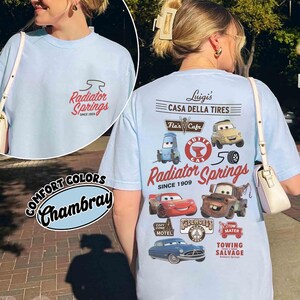 May include: A light blue t-shirt featuring a small Radiator Springs logo on the front and a large graphic on the back with characters and landmarks from the movie Cars, including Luigi's Casa Della Tires, Flo's Cafe, and Tow Mater Towing Salvage.