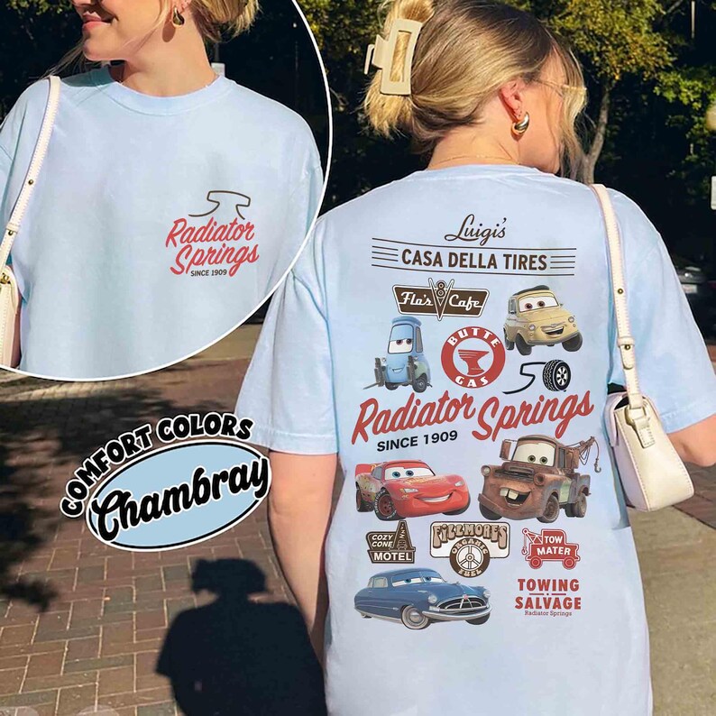 May include: A light blue t-shirt featuring a small Radiator Springs logo on the front and a large graphic on the back with characters and landmarks from the movie Cars, including Luigi's Casa Della Tires, Flo's Cafe, and Tow Mater Towing Salvage.