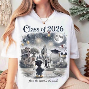 May include: White t-shirt featuring a colorful illustration with the text "Class of 2026" and "from the tassel to the castle." The graphic includes a graduation Mickey Mouse, fireworks, and iconic landmarks.