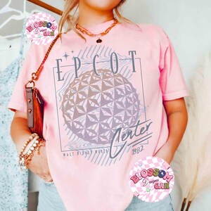 May include: Pink t-shirt featuring a graphic of the EPCOT Center sphere with the text "EPCOT Center 1982" in a retro style. The design includes lines and stars, with "Walt Disney World" below. The shirt is a light pink color.