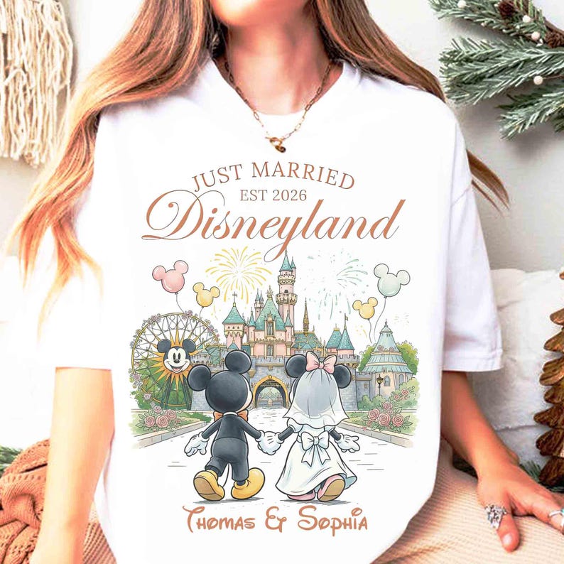 Może przedstawiać: Biała koszulka z grafiką Disneylandu przedstawiającą Mickey i Minnie Mouse jako parę młodą, trzymających się za ręce przed zamkiem. Tekst brzmi "Just Married Disneyland EST 2026" oraz imiona "Thomas & Sophia".