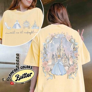May include: Butter yellow t-shirt with a Cinderella theme. The front features a high heel, dress, carriage, and castle with the text "meet me at midnight." The back has a castle scene with Cinderella in a floral frame.