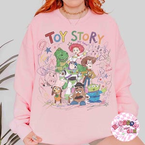 Puede incluir: Sudadera rosa claro con un gr&aacute;fico colorido de personajes de Toy Story y la frase "TOY STORY". El dise&ntilde;o incluye a Woody, Buzz Lightyear y otros personajes de la pel&iacute;cula, con acentos garabateados.
