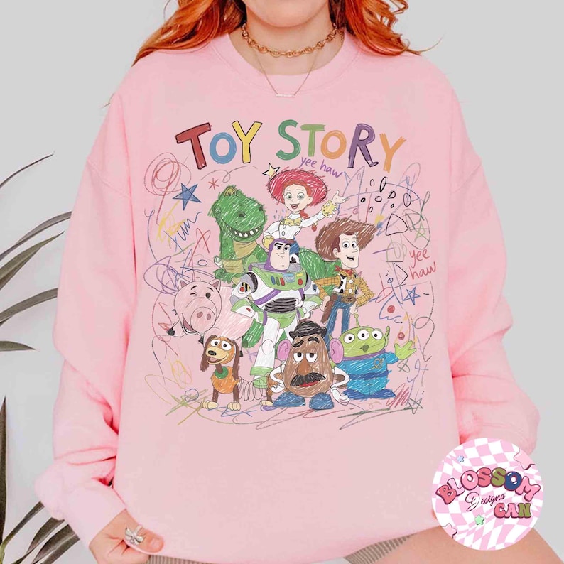 Puede incluir: Sudadera rosa claro con un gr&aacute;fico colorido de personajes de Toy Story y la frase "TOY STORY". El dise&ntilde;o incluye a Woody, Buzz Lightyear y otros personajes de la pel&iacute;cula, con acentos garabateados.