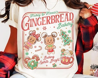 May include: Cream-colored t-shirt featuring a festive design. The graphic includes the text "Mickey & Minnie's Gingerbread Bakery" with gingerbread characters, a Christmas tree, donuts, and a mug of hot cocoa. The phrase "Baking Spirits Bright" is also included.