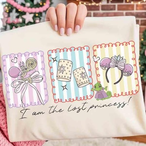Op de afbeelding: Crèmekleurig T-shirt met drie geïllustreerde panelen met een prinsessenthema. De panelen bevatten een lolly, lantaarns en muisoren. De tekst "I am the lost princess!" staat eronder.