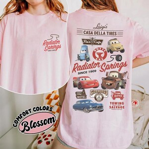 May include: A light pink t-shirt featuring a small Radiator Springs logo on the front. The back displays a large graphic with various Cars characters, including Lightning McQueen and Mater, alongside text like Luigi's Casa Della Tires and Flo's Cafe.