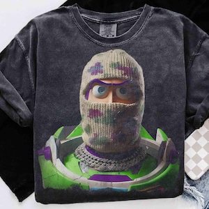 May include: A charcoal gray short-sleeve t-shirt featuring a graphic print of a character wearing a beige knit balaclava with purple and green accents. The character has large blue eyes and wears a silver chain necklace over a green space suit.