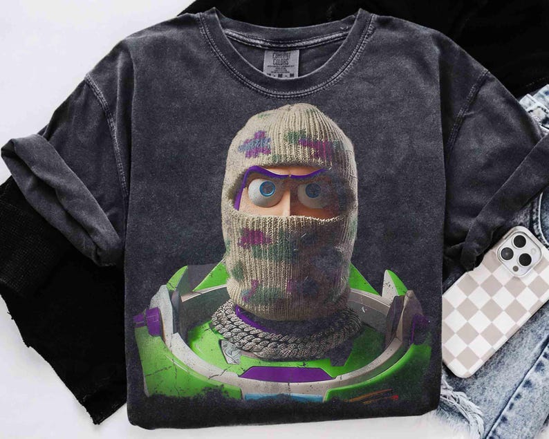 May include: A charcoal gray short-sleeve t-shirt featuring a graphic print of a character wearing a beige knit balaclava with purple and green accents. The character has large blue eyes and wears a silver chain necklace over a green space suit.