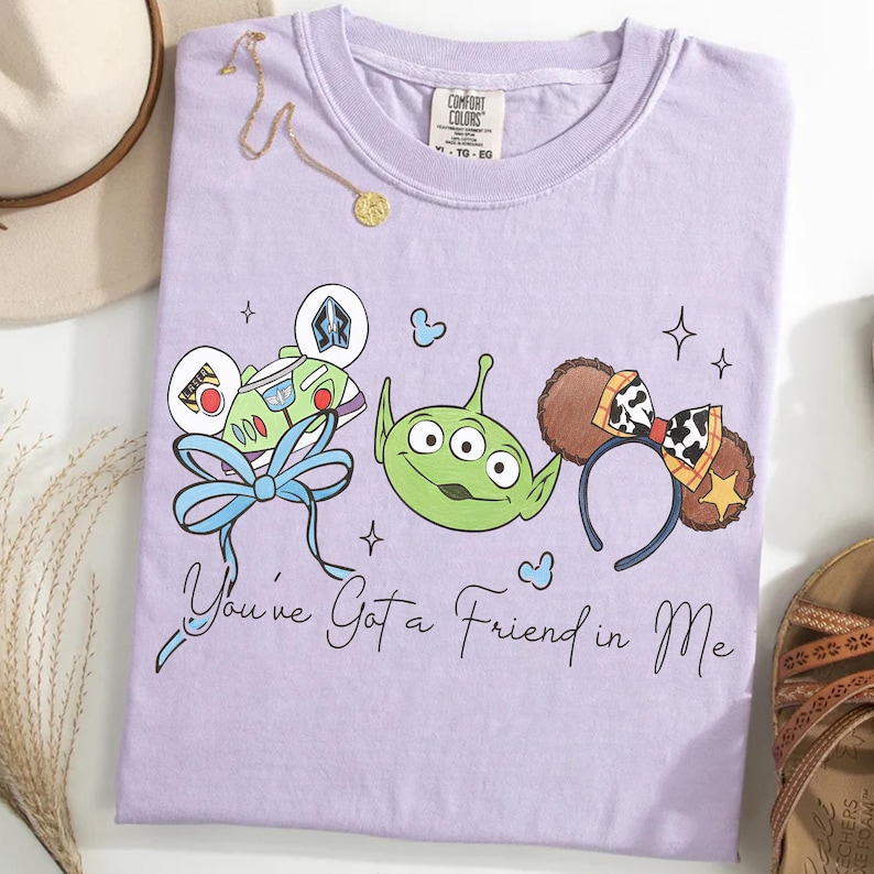 May include: A lilac Comfort Colors t-shirt featuring cartoon characters and the text "You've Got a Friend in Me." The design includes Buzz Lightyear, a green alien, and Mickey Mouse ears with a cow print bow.