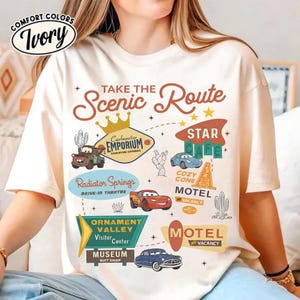 May include: Ivory-colored t-shirt with a graphic design featuring the text "Take the Scenic Route" and illustrations of cars and roadside attractions. The design includes the words "Radiator Springs" and "Motel".