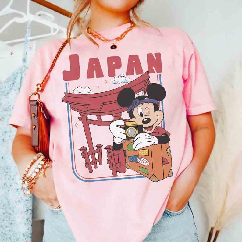 Pode incluir: Camiseta rosa claro com um desenho do Mickey Mouse tirando uma foto em frente a um port&atilde;o Torii japon&ecirc;s. A palavra "JAPAN" est&aacute; impressa acima do desenho. O design inclui nuvens e uma mala.
