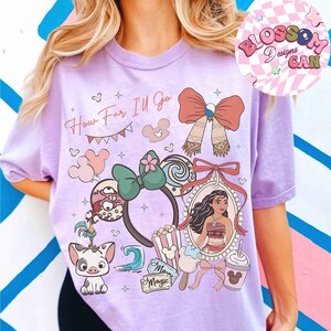 May include: A light purple t-shirt featuring a colorful graphic design. The design includes the text "How Far I'll Go", a portrait, and various cartoon elements. The shirt has short sleeves and a round neckline.