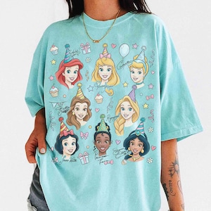May include: A light blue t-shirt featuring illustrations of several Disney princesses wearing party hats. The shirt is decorated with cupcakes, stars, bows, and balloons. Each princess has a handwritten name below her portrait.