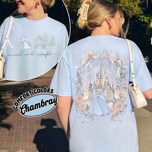 May include: Light blue Comfort Colors Chambray t-shirt featuring a Cinderella theme. The front has a shoe, dress, carriage, and castle with "meet me at midnight" text. The back has a castle, Cinderella, and floral design.
