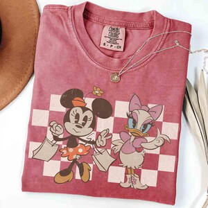May include: A dusty rose Comfort Colors t-shirt featuring Minnie Mouse and Daisy Duck with shopping bags, set against a checkered background. The shirt has a crew neck and short sleeves. A silver necklace with a pendant is draped across the shirt.