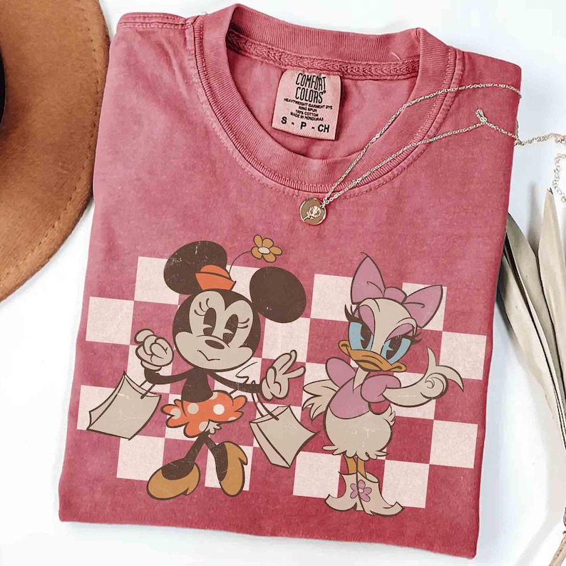 May include: A dusty rose Comfort Colors t-shirt featuring Minnie Mouse and Daisy Duck with shopping bags, set against a checkered background. The shirt has a crew neck and short sleeves. A silver necklace with a pendant is draped across the shirt.