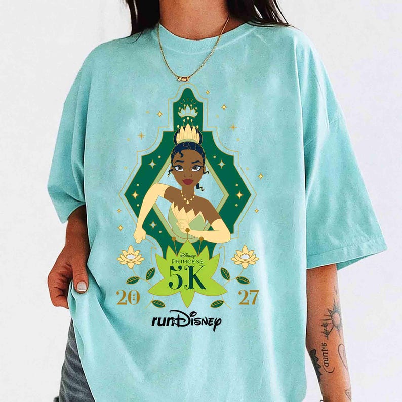May include: Light blue t-shirt featuring a graphic of Princess Tiana from Disney. The design includes a green and gold geometric frame, the text "Disney Princess 5K", and the "runDisney" logo. The numbers "20" and "27" are also present.