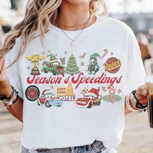May include: White t-shirt featuring a colorful graphic with characters from the movie Cars, including Mater, Lightning McQueen, and others, in festive holiday attire. The text "Season's Speedings" is displayed in red script.