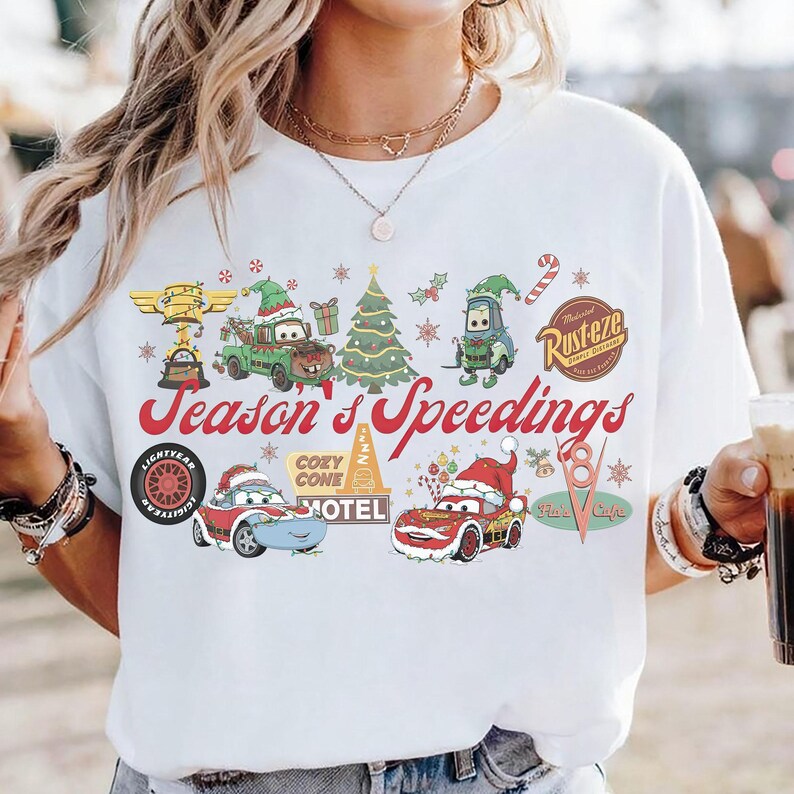 May include: White t-shirt featuring a colorful graphic with characters from the movie Cars, including Mater, Lightning McQueen, and others, in festive holiday attire. The text "Season's Speedings" is displayed in red script.