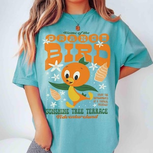 May include: A turquoise t-shirt featuring the Orange Bird character with text that reads "Home of the Orange Bird" in orange. The shirt also includes the text "Sunshine Tree Terrace Adventureland" and "Enjoy the refreshments of a tropical paradise!"
