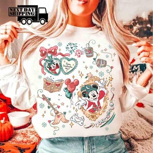 Könnte beinhalten: Weißes Sweatshirt mit einem farbenfrohen Mickey Mouse-Design. Die Grafik zeigt Mickey in einem Zaubererkostüm, ein Buch, Luftballons und andere verspielte Elemente. Das Wort "FANTASY" ist ebenfalls sichtbar.