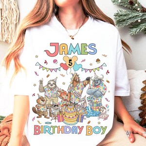 K&ouml;nnte beinhalten: Wei&szlig;es T-Shirt mit einem farbenfrohen Grafikdesign f&uuml;r einen Geburtstag. Das Design enth&auml;lt den Namen "JAMES", die Zahl "5" und die Worte "BIRTHDAY BOY". Die Grafik zeigt Zeichentricktiere und eine Geburtstagstorte.