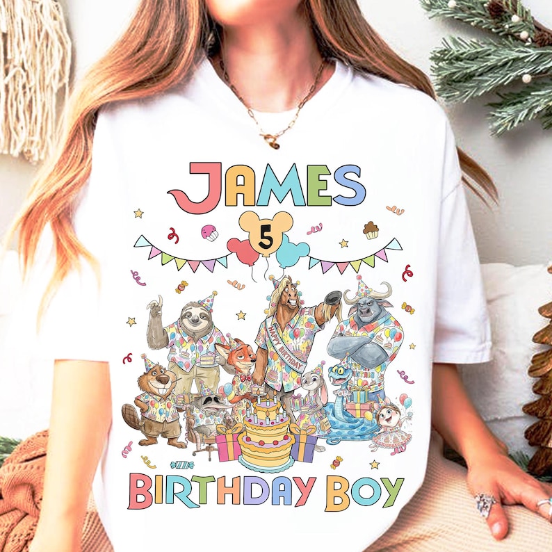 K&ouml;nnte beinhalten: Wei&szlig;es T-Shirt mit einem farbenfrohen Grafikdesign f&uuml;r einen Geburtstag. Das Design enth&auml;lt den Namen "JAMES", die Zahl "5" und die Worte "BIRTHDAY BOY". Die Grafik zeigt Zeichentricktiere und eine Geburtstagstorte.