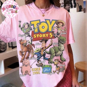 May include: A light pink t-shirt featuring a colorful graphic print of Toy Story characters including Buzz, Jessie, Bullseye, Alien, Woody, Rex, Hamm, and Slinky. The text reads Toy Story 5, Since 1995, and Hi! with character names labeled.