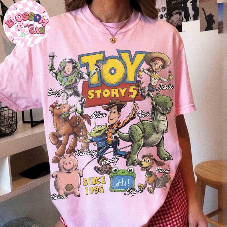 May include: A light pink t-shirt featuring a colorful graphic print of Toy Story characters including Buzz, Jessie, Bullseye, Alien, Woody, Rex, Hamm, and Slinky. The text reads Toy Story 5, Since 1995, and Hi! with character names labeled.