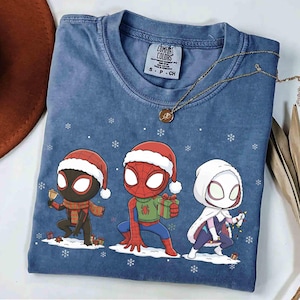 May include: Blue t-shirt with three Spider-Man characters in Santa hats, holding gifts and Christmas lights. The shirt has a Comfort Colors tag and a gold necklace. Snowflakes are scattered around the characters.