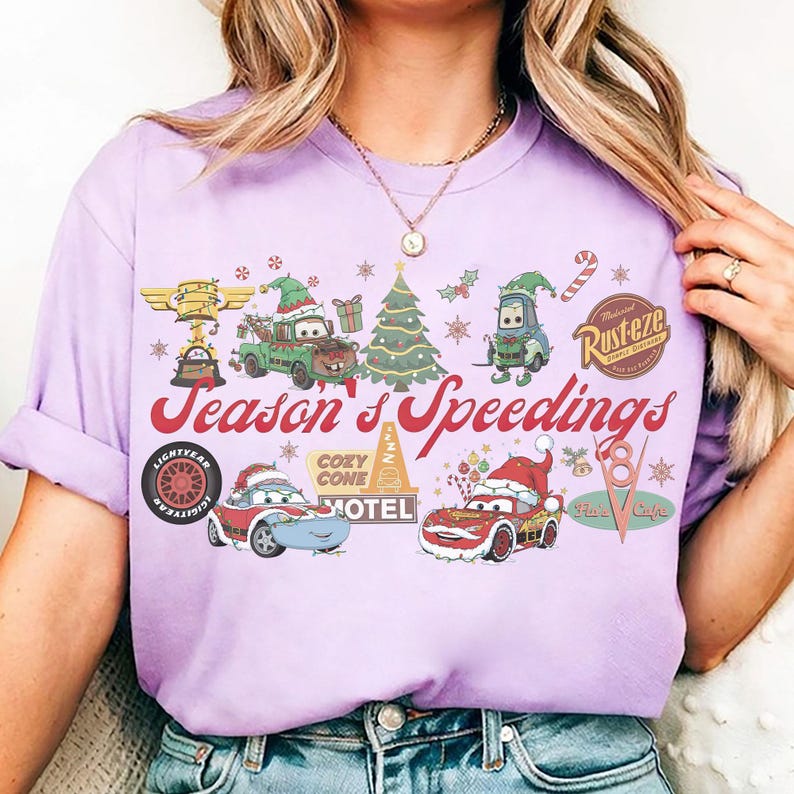 May include: A light purple t-shirt featuring a festive graphic with characters from the movie Cars. The design includes Christmas elements, the text "Season's Speedings", and various Cars-themed logos and illustrations.