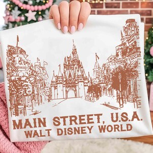 Peut inclure: Tissu blanc avec une illustration marron de Main Street, U.S.A. &agrave; Walt Disney World. L'image pr&eacute;sente des dessins d&eacute;taill&eacute;s de b&acirc;timents, un ch&acirc;teau et le texte "MAIN STREET, U.S.A. WALT DISNEY WORLD" en marron.