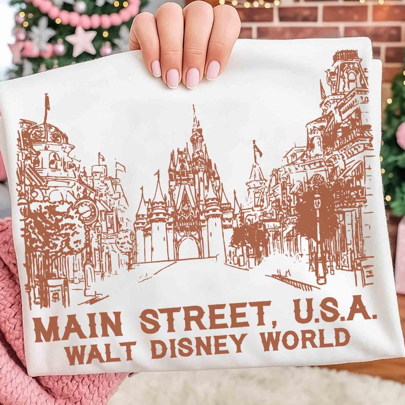 Peut inclure: Tissu blanc avec une illustration marron de Main Street, U.S.A. &agrave; Walt Disney World. L'image pr&eacute;sente des dessins d&eacute;taill&eacute;s de b&acirc;timents, un ch&acirc;teau et le texte "MAIN STREET, U.S.A. WALT DISNEY WORLD" en marron.