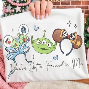 May include: White t-shirt featuring cartoon illustrations of Buzz Lightyear, an alien, and a headband with brown ears and a cow print bow. The text "You've Got a Friend in Me" is written in cursive below the illustrations.