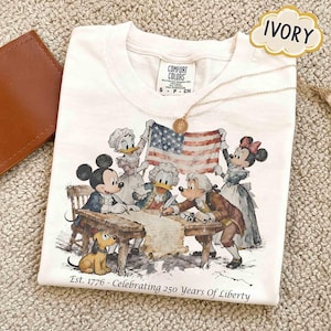 May include: Ivory-colored t-shirt featuring a graphic of Disney characters dressed in historical attire, holding an American flag and signing a document. The text "Est. 1776 - Celebrating 250 Years Of Liberty" is printed below the graphic. A gold necklace is draped on the shirt.