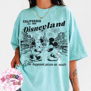 May include: A light blue t-shirt featuring a graphic of Mickey and Minnie Mouse in front of a Disneyland scene. The shirt includes the text "CALIFORNIA Est. 1955 Disneyland" and "The happiest place on earth."